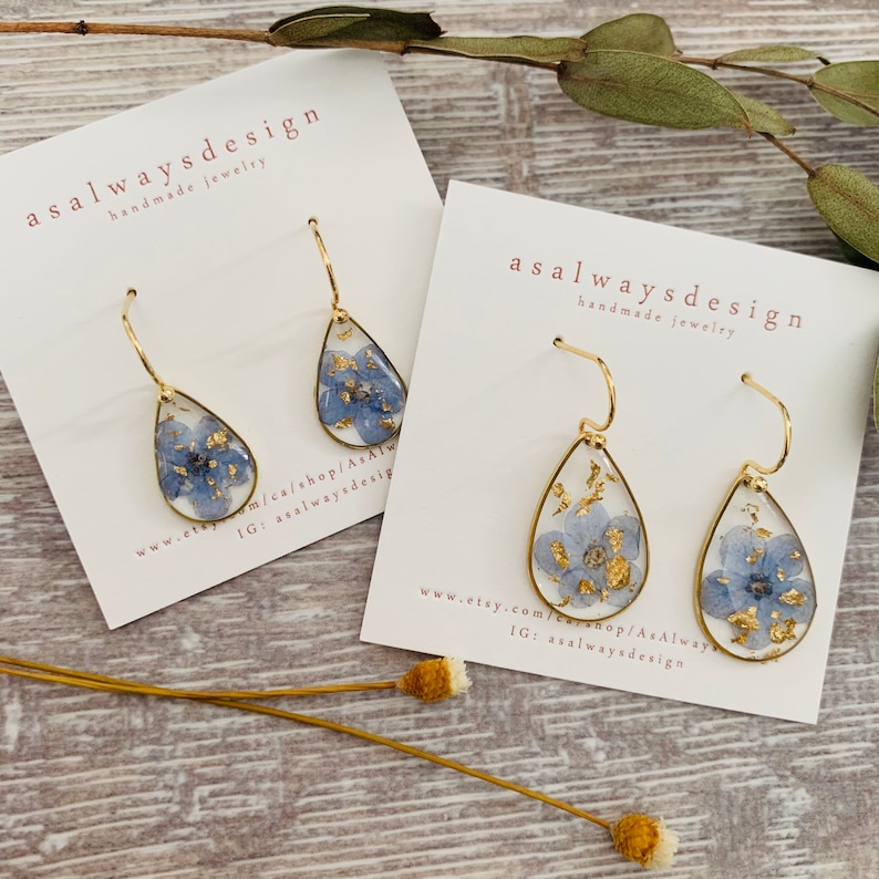 May include: A pair of gold teardrop earrings with blue pressed flowers and gold leaf accents. The earrings are on white cards with the text "asalwaysdesign handmade jewelry" and website information.