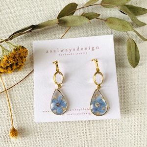 May include: Gold earrings with a teardrop-shaped resin pendant featuring a pressed blue flower and gold flecks. The earrings also feature a clear round crystal charm.