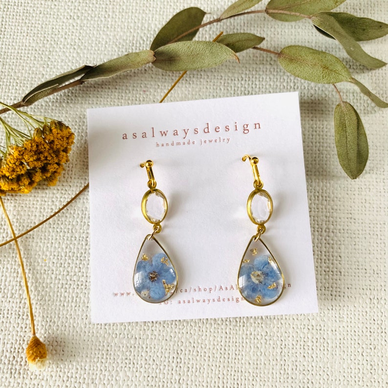 May include: Gold earrings with a teardrop-shaped resin pendant featuring a pressed blue flower and gold flecks. The earrings also feature a clear round crystal charm.