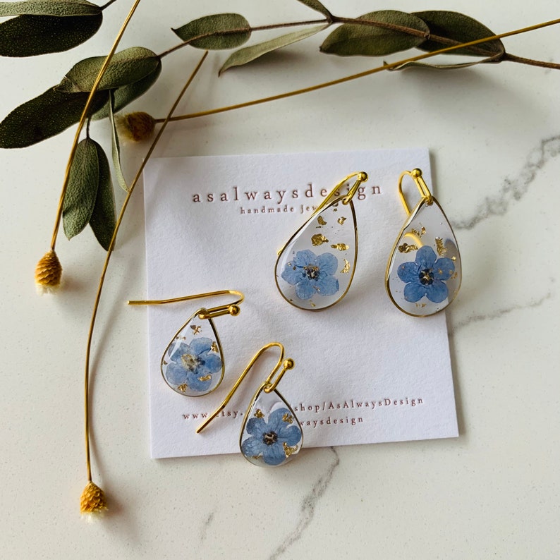 May include: A set of three gold teardrop earrings with blue pressed flowers and gold leaf accents. The earrings are displayed on a white card with the text "asalwaysdesign handmade jewelry" and "shop/AsAlwaysDesign waysdesign".