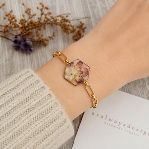 May include: A gold chain bracelet featuring a central hexagonal pendant containing pressed white and purple flowers with gold leaf accents. The bracelet is secured with a gold link chain and is worn on a wrist covered by a beige knit sleeve.