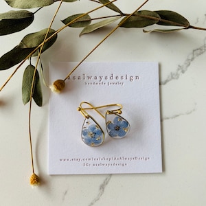 May include: A pair of gold teardrop earrings with a blue forget-me-not flower pressed inside each earring. The earrings are on a white card with the text "asalwaysdesign handmade jewelry" and website information.