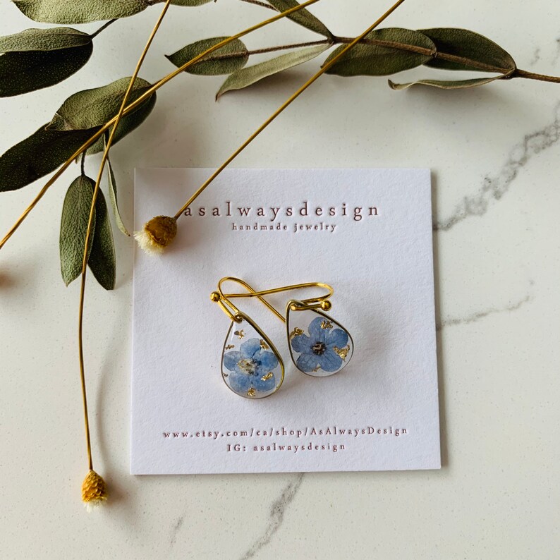 May include: A pair of gold teardrop earrings with a blue forget-me-not flower pressed inside each earring. The earrings are on a white card with the text "asalwaysdesign handmade jewelry" and website information.