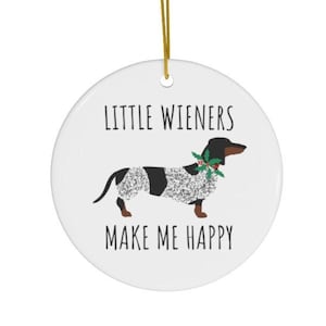 May include: A white ceramic ornament with a dachshund illustration. The dog is black and white with a green bow. The text "LITTLE WIENERS MAKE ME HAPPY" is printed on the ornament, which hangs from a gold string.