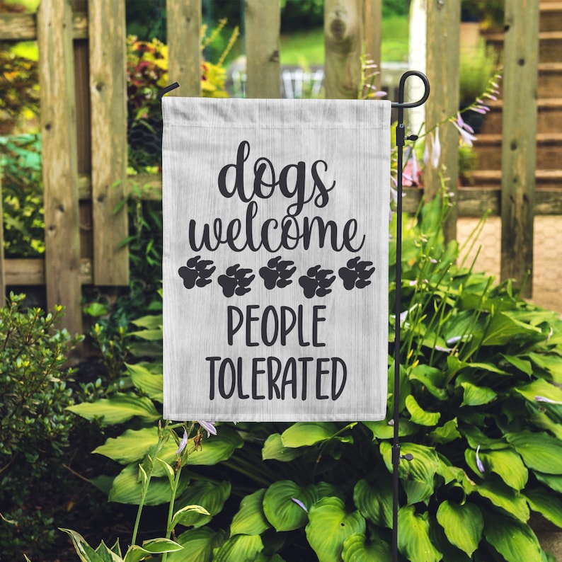 May include: White garden flag with black text that reads "dogs welcome" with paw prints and "people tolerated".
