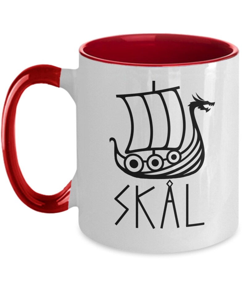 May include: White ceramic coffee mug with a red rim and handle. The mug features a black illustration of a Viking ship with a dragon head and the word "SKAL" written in runes.
