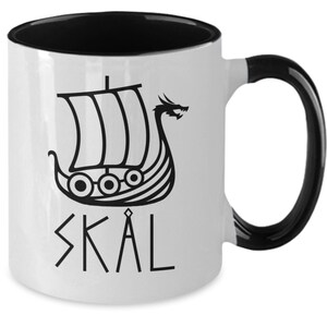 May include: White ceramic mug with a black rim and handle. The mug features a black and white illustration of a Viking ship with a dragon head and the word "SK&Aring;L" in black lettering.