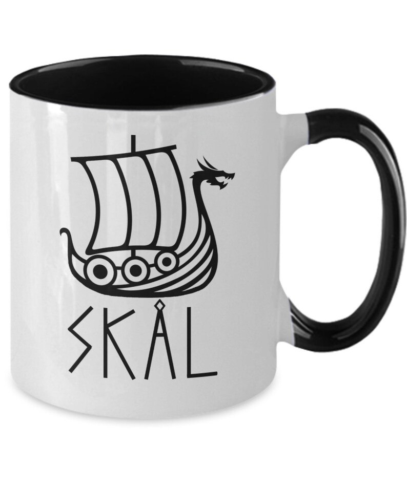 May include: White ceramic mug with a black rim and handle. The mug features a black and white illustration of a Viking ship with a dragon head and the word "SK&Aring;L" in black lettering.