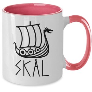 May include: White ceramic coffee mug with a pink interior and handle. The mug features a black and white illustration of a Viking ship with a dragon head and the word "SKAL" in black.