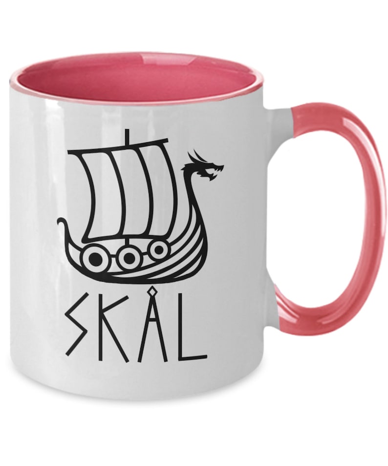 May include: White ceramic coffee mug with a pink interior and handle. The mug features a black and white illustration of a Viking ship with a dragon head and the word "SKAL" in black.