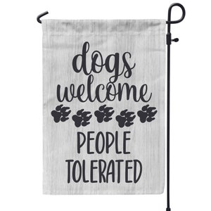 May include: A white garden flag with a woodgrain pattern and black text that reads "dogs welcome people tolerated" with five black paw prints below the text.