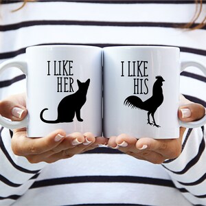 May include: Two white ceramic mugs with black text and illustrations. One mug says "I LIKE HER" with a black cat silhouette. The other says "I LIKE HIS" with a rooster silhouette. The mugs are held by a person.