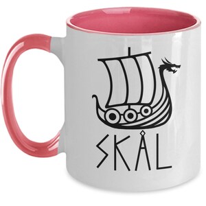 May include: White ceramic coffee mug with a pink rim and handle. The mug features a black silhouette of a Viking ship with a dragon head and the word "SKAL" written in runes.