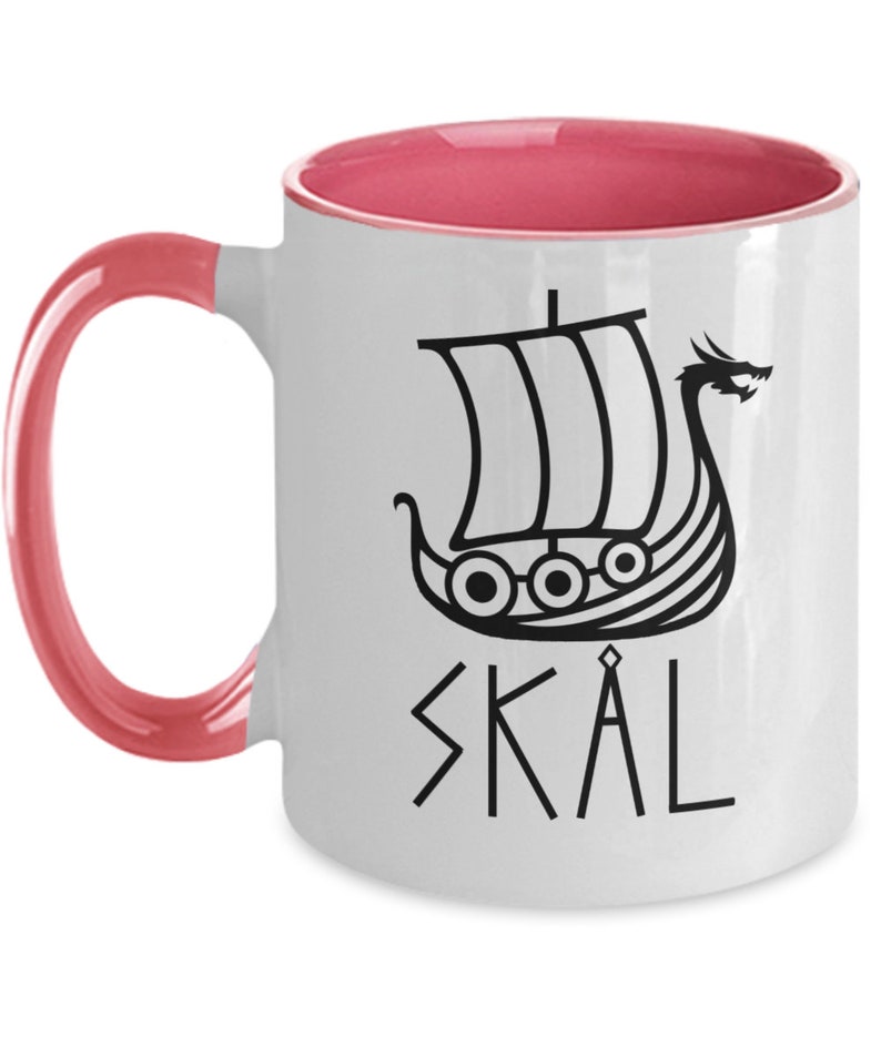 May include: White ceramic coffee mug with a pink rim and handle. The mug features a black silhouette of a Viking ship with a dragon head and the word "SKAL" written in runes.