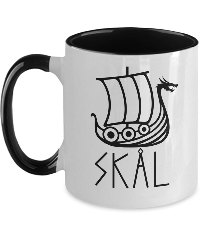 May include: White ceramic coffee mug with a black interior and handle. The mug features a black and white illustration of a Viking ship with a dragon head and the word "SKAL" written in runes.