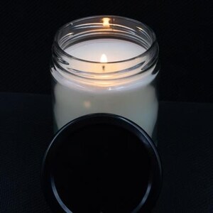 May include: A clear glass jar candle with a white wax and a black lid. The candle is lit and the flame is visible.