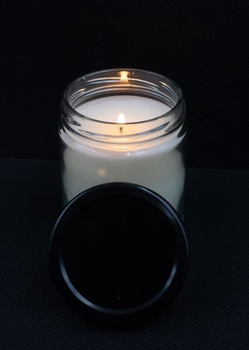 May include: A clear glass jar candle with a white wax and a black lid. The candle is lit and the flame is visible.