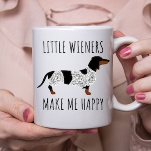 K&ouml;nnte beinhalten: Wei&szlig;e Keramik-Kaffeetasse mit einem schwarzen Dackel, der einen glitzernden wei&szlig;en und schwarzen Mantel tr&auml;gt. Die Tasse tr&auml;gt die Aufschrift "Little Wieners Make Me Happy."