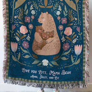 May include: A teal woven throw blanket with a bear family illustration and floral design. The blanket has a fringed edge and the text "Love You Lots, Mama Bear" with names below. The blanket is held up by a person.