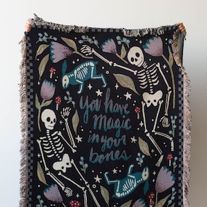 May include: A black woven throw blanket with a whimsical design featuring skeletons, flowers, and mushrooms. The text "You have magic in your bones" is written in a stylized font.