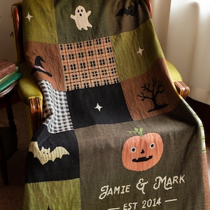 May include: A woven throw blanket featuring a patchwork design with Halloween motifs including ghosts, bats, pumpkins, a witch hat, and a black cat. The blanket displays the text "JAMIE & MARK" and "EST 2014" in white, finished with a multi-colored fringe edge.
