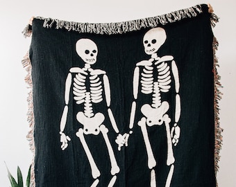 May include: A black woven throw blanket with two white skeleton figures holding hands. The blanket has fringe around the edges.