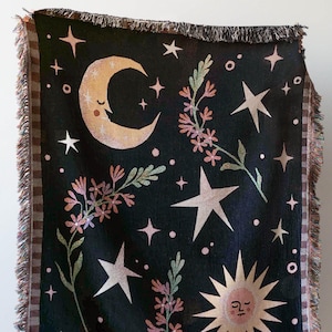 Peut inclure: Une couverture en tissage noir avec un motif fantaisiste représentant une lune croissante, un soleil, des étoiles et des brins de fleurs en rose, vert et jaune.