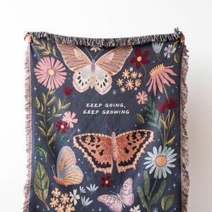 Woven blanket being held up by a person. Features a floral and butterfly design and the words "keep going, keep growing"
