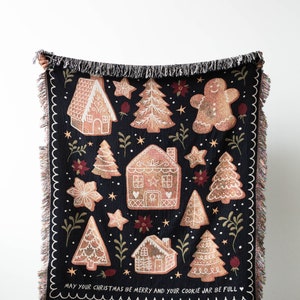 K&ouml;nnte beinhalten: Eine schwarze gewebte Wolldecke mit einem Lebkuchen-Motiv. Die Decke zeigt Lebkuchenh&auml;user, B&auml;ume und Sterne. Der Text "May your Christmas be merry and your cookie jar be full" ist unten auf der Decke aufgedruckt.