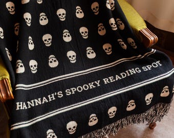 May include: A black woven throw blanket featuring a repeating pattern of off-white skulls. The blanket includes a horizontal border with the text "HANNAH'S SPOOKY READING SPOT" in off-white lettering and finished with a fringed edge at the bottom.