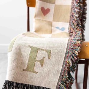 May include: A woven throw blanket with a large letter "F" in the centre. The blanket has a checkered pattern with hearts in each square. The blanket has black fringe on the bottom and multicoloured fringe on the sides.