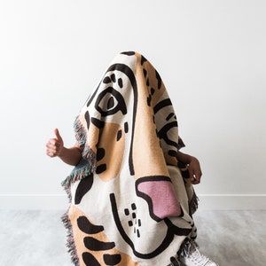 May include: A person is sitting in a chair, covered in a colourful woven blanket with a large abstract design. The blanket is mostly beige and white with black and pink accents. The person is wearing white trainers.