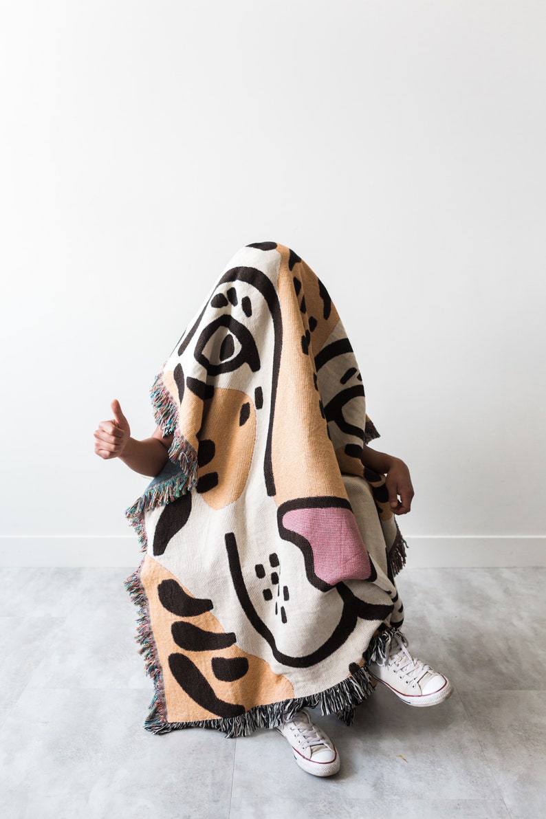 May include: A person is sitting in a chair, covered in a colourful woven blanket with a large abstract design. The blanket is mostly beige and white with black and pink accents. The person is wearing white trainers.