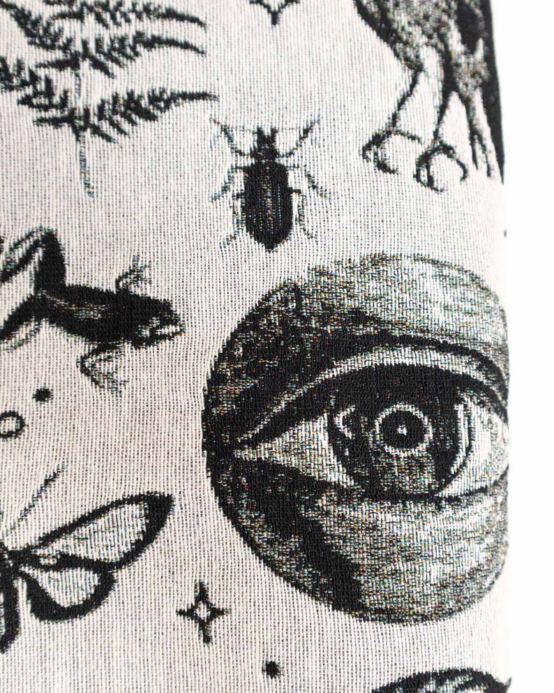 May include: A woven textile fabric featuring a black and white pattern of a large human eye, a beetle, a frog, a moth, and fern leaves. The design is rendered in a high-contrast monochrome style with intricate details on each of the natural elements.