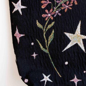 May include: Black fabric with a floral and star pattern. The fabric has a textured, woven look. The flowers are pink, purple, and green. The stars are white and silver.