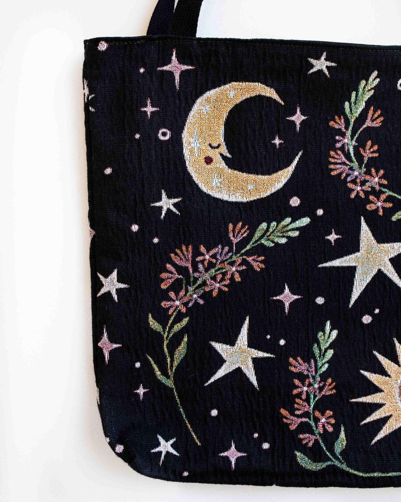 May include: Black tote bag with a moon, stars, and floral design. The moon has a smiling face and the stars are white. The flowers are pink, green, and orange.