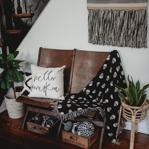 May include: A wooden bench with a black and white skull patterned throw blanket and a white pillow with the text "hello pumpkin" in black lettering. The bench is decorated with pumpkins and is in front of a staircase with a black and white woven wall hanging.