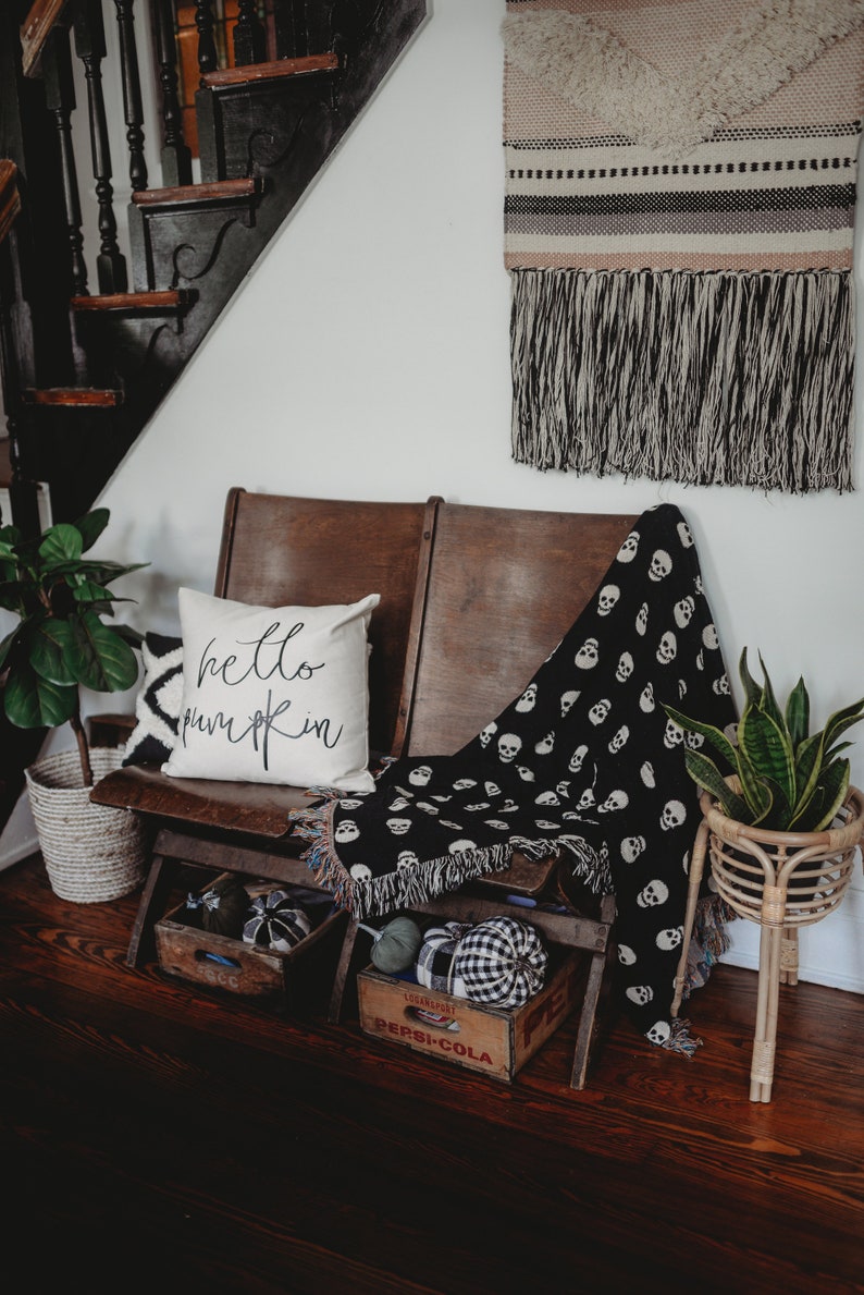 May include: A wooden bench with a black and white skull patterned throw blanket and a white pillow with the text "hello pumpkin" in black lettering. The bench is decorated with pumpkins and is in front of a staircase with a black and white woven wall hanging.