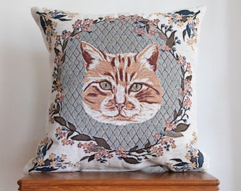 May include: A cream-colored throw pillow with a floral design and a brown and orange cat embroidered in the center.