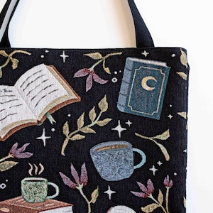 May include: Black tote bag with a tapestry design featuring books, coffee cups, stars, and leaves. The bag has a black strap.