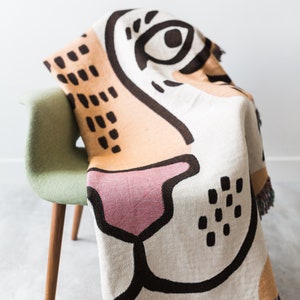 May include: A woven throw blanket with a colourful abstract design featuring a large eye, nose, and mouth. The blanket is draped over a green chair with wooden legs.