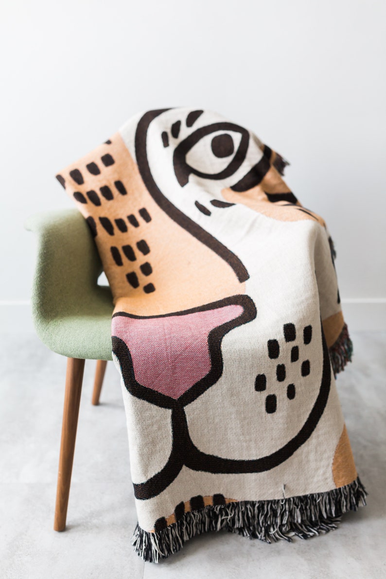 May include: A woven throw blanket with a colourful abstract design featuring a large eye, nose, and mouth. The blanket is draped over a green chair with wooden legs.