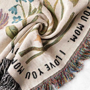 May include: Cream-colored woven throw blanket with a floral design in shades of blue, green, and yellow. The blanket has the text "I LOVE YOU MOM" in black and a fringed edge with multicolored threads.