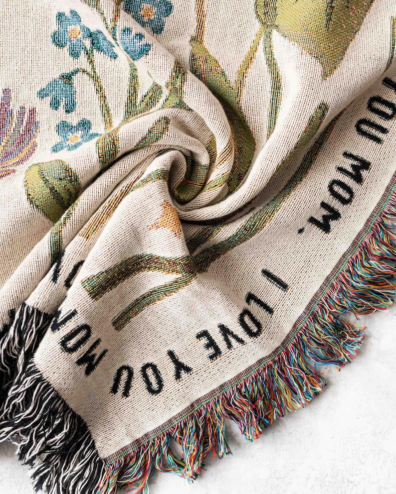 May include: Cream-colored woven throw blanket with a floral design in shades of blue, green, and yellow. The blanket has the text "I LOVE YOU MOM" in black and a fringed edge with multicolored threads.