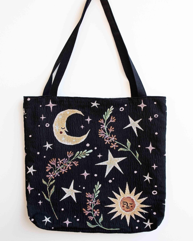 May include: Black tote bag with a celestial design featuring a crescent moon, a sun with a face, and stars. The bag has black straps.