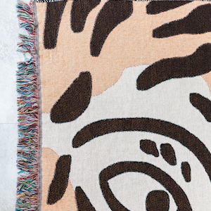 May include: A woven throw blanket with an abstract black and white pattern on a peach background. The blanket has a fringe border with multicoloured threads.