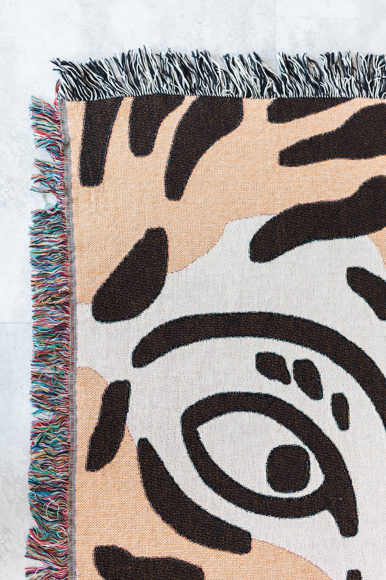 May include: A woven throw blanket with an abstract black and white pattern on a peach background. The blanket has a fringe border with multicoloured threads.