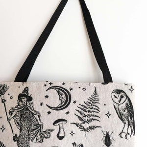 May include: A beige tote bag with black woven straps featuring a black ink-style print of a witch holding a broom, a crescent moon, stars, a mushroom, a fern leaf, an owl, a beetle, a frog, and an open book on a light-colored fabric surface.