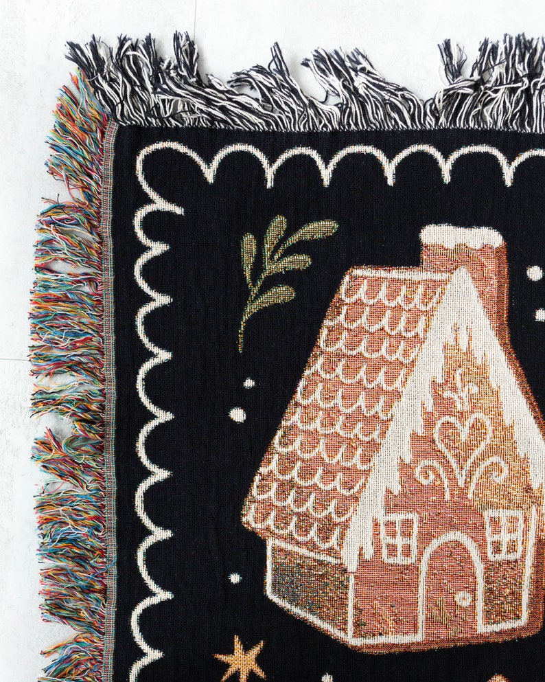 K&ouml;nnte beinhalten: Eine schwarze gewebte Wandteppich mit einem Lebkuchenhaus-Design. Das Haus ist braun mit wei&szlig;em Zuckerguss und einem roten Dach. Auf dem schwarzen Hintergrund befinden sich goldene Sterne und ein gr&uuml;ner Zweig.
