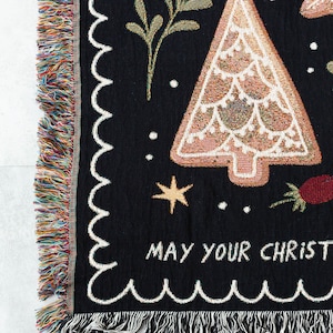 K&ouml;nnte beinhalten: Schwarze gewebte Wurfdecke mit einem Lebkuchenbaum-Design und dem Text "May your Christmas be merry and bright". Die Decke hat einen gewellten Rand und Fransen.